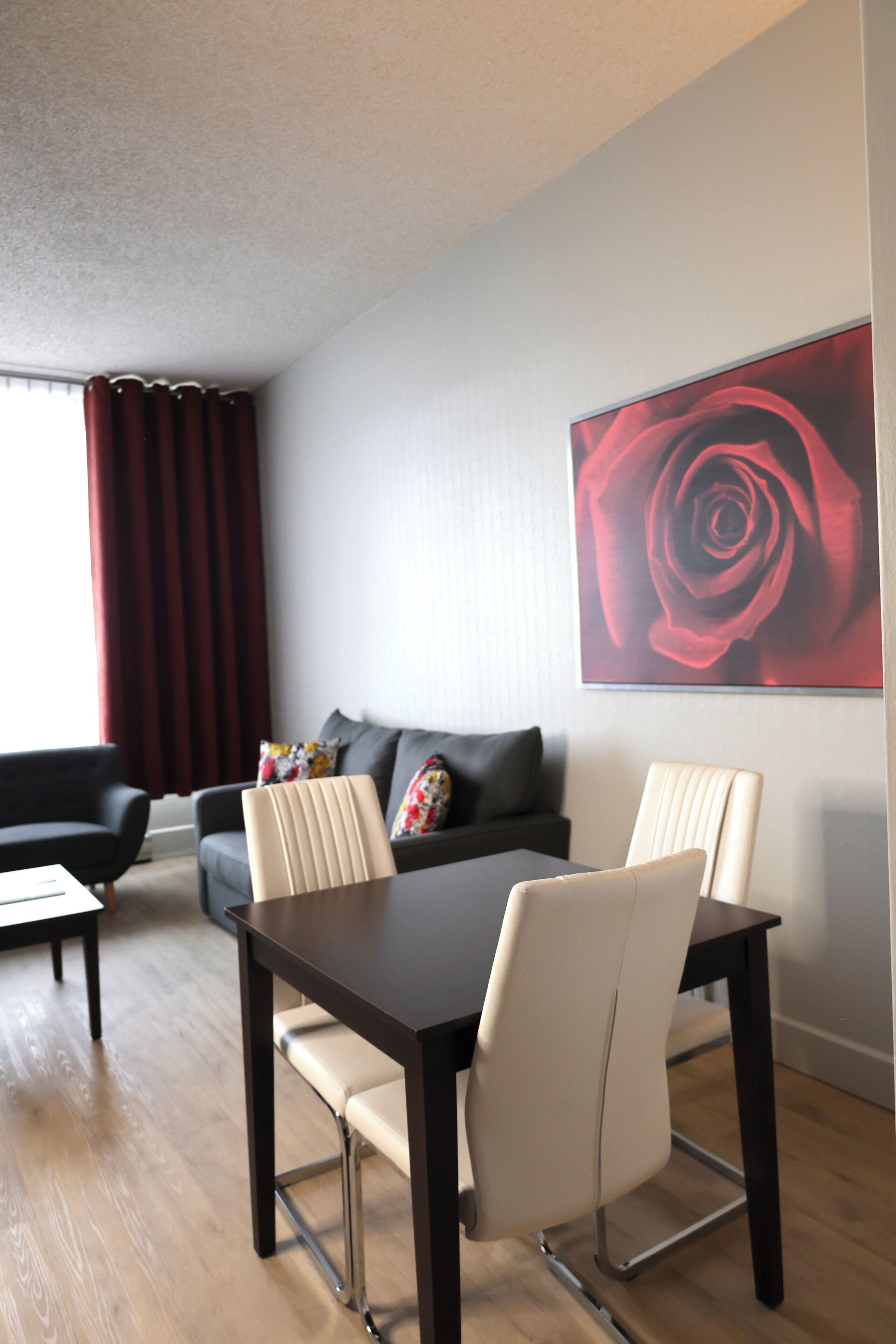 A modern living room with a dining table, two chairs, a sofa, and a large red rose painting on the wall.