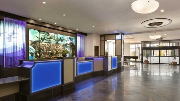A hotel reception area with a modern design, featuring a reception desk with blue lighting, a large window with a view of greenery, and a spacious lobby with a chandelier. - Travelodge by Wyndham Hotel & Convention Centre Quebec City (Québec City)