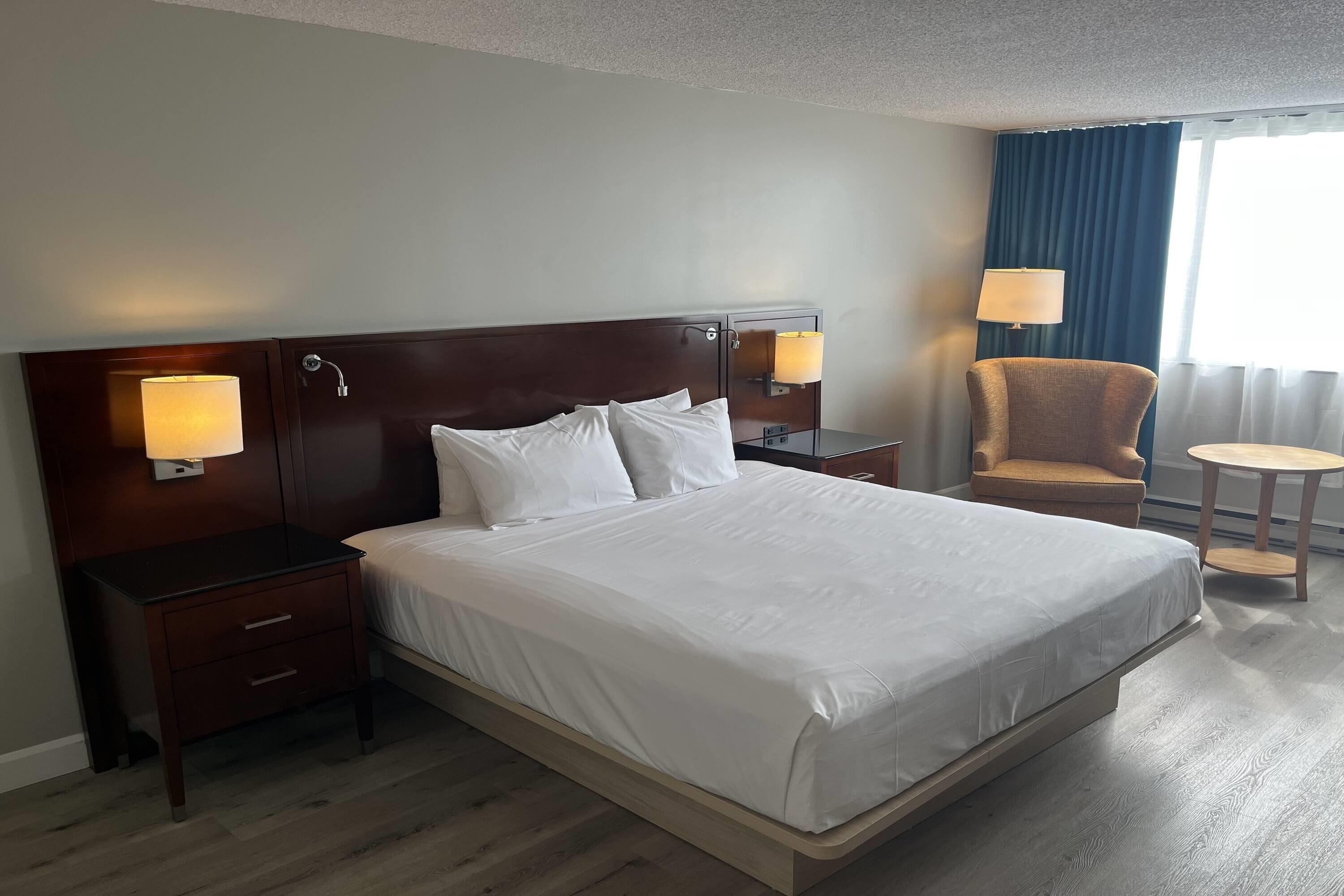 Habitación de hotel con una cama grande, lámparas de noche, una silla, una mesita y un ventanal con cortinas.