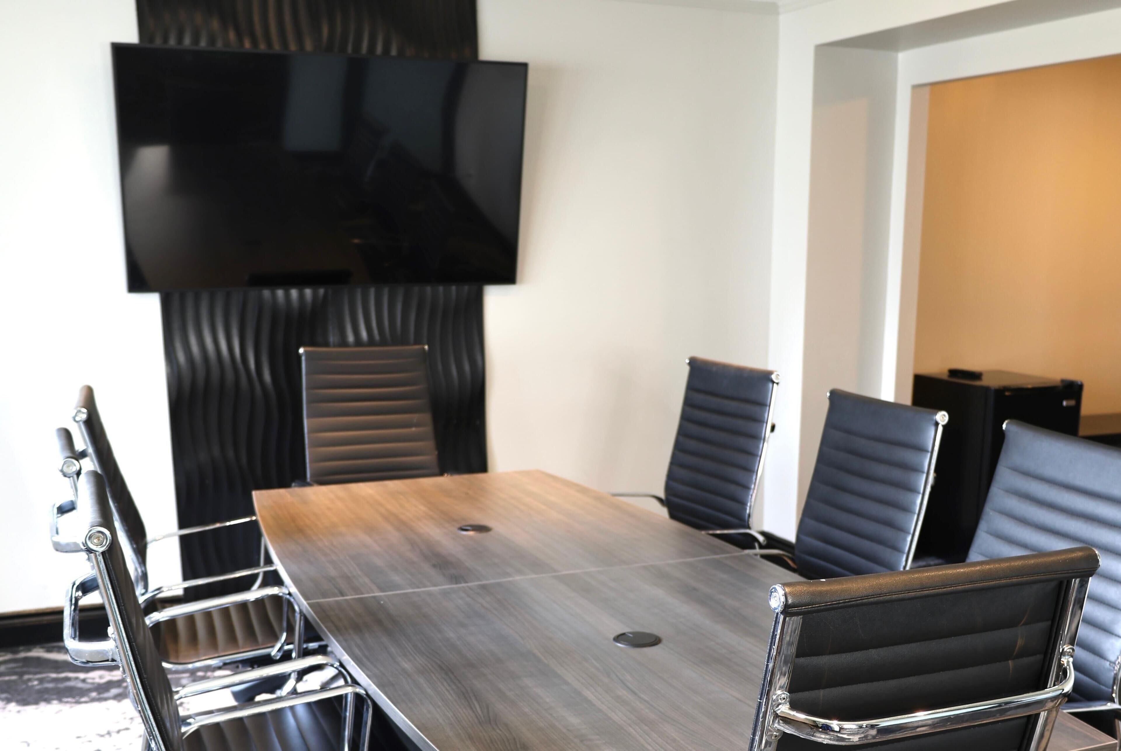 Une salle de conférence avec une longue table, six chaises, un téléviseur à écran plat fixé au mur et une porte donnant sur une autre pièce.