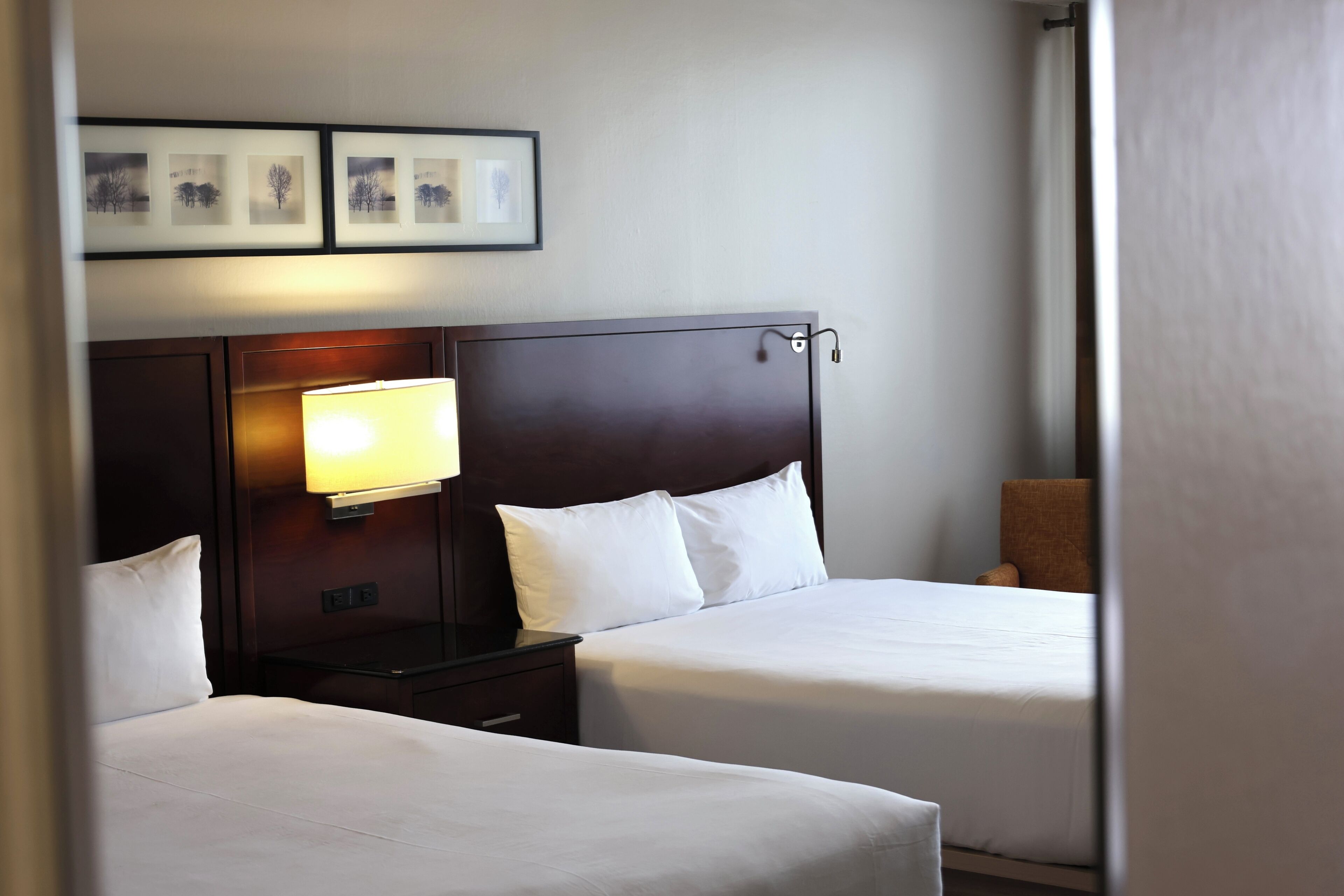 A hotel room with two beds, a nightstand, a lamp, and a wall-mounted picture.