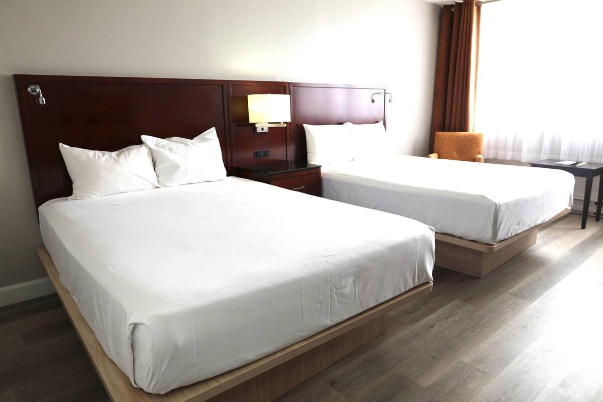 Habitación de hotel con dos camas, cabeceros de madera, mesitas de noche con lámparas y vistas al exterior.