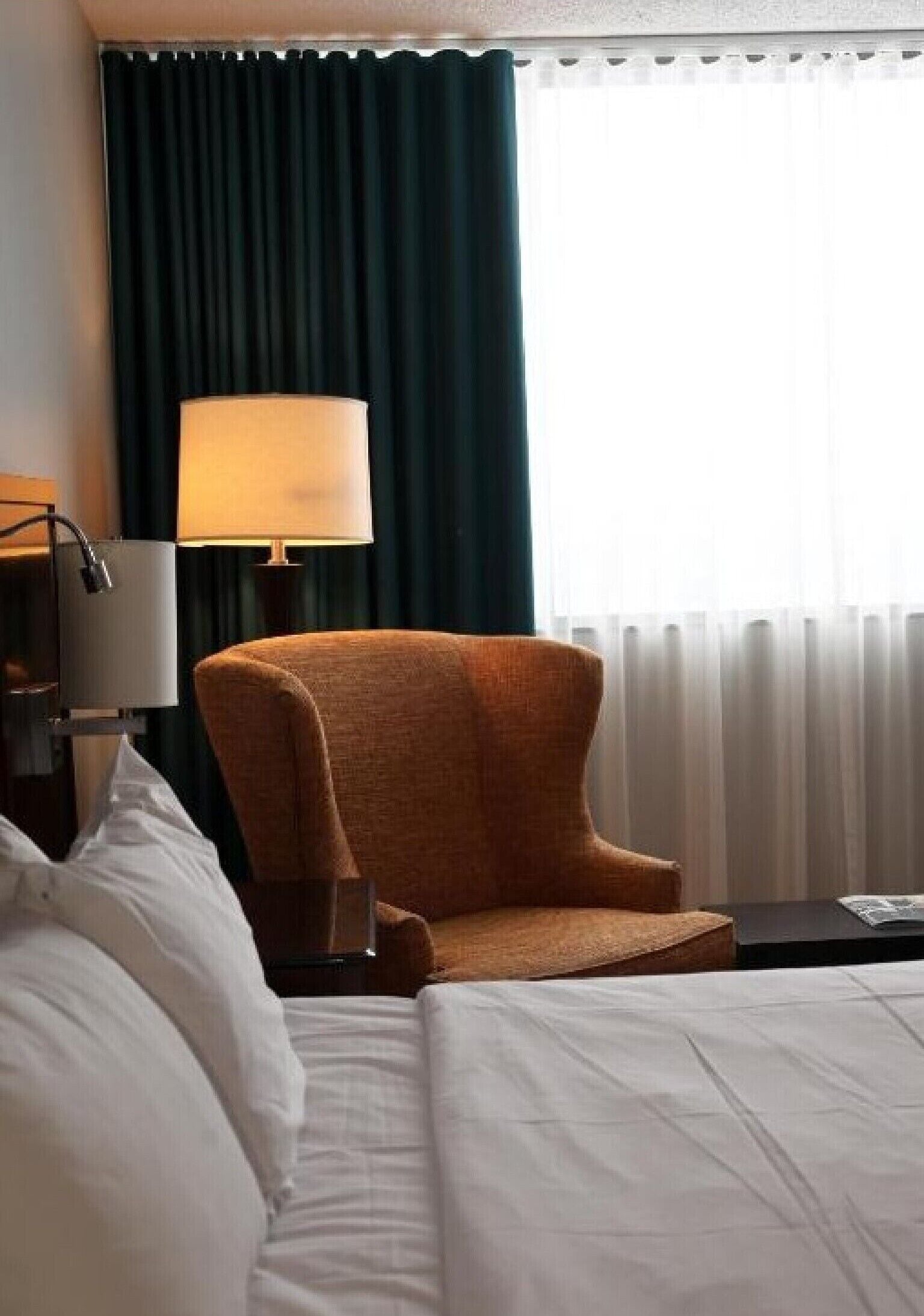 A hotel room with a bed, a chair, a lamp, a nightstand, and a window with curtains.