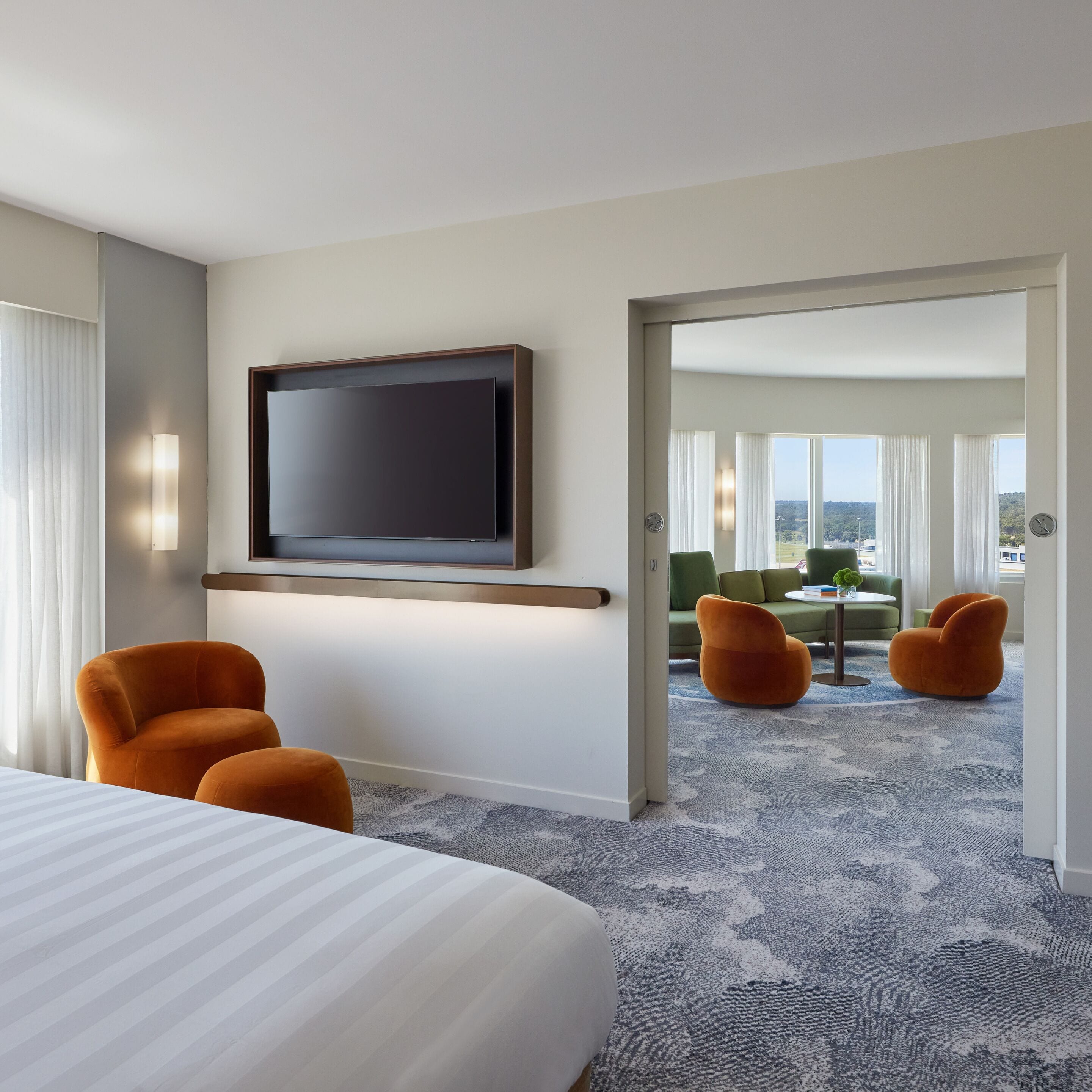 A hotel room with a large bed, two orange armchairs, a flat-screen TV mounted on the wall, and a view of a green outdoor area through an open door.