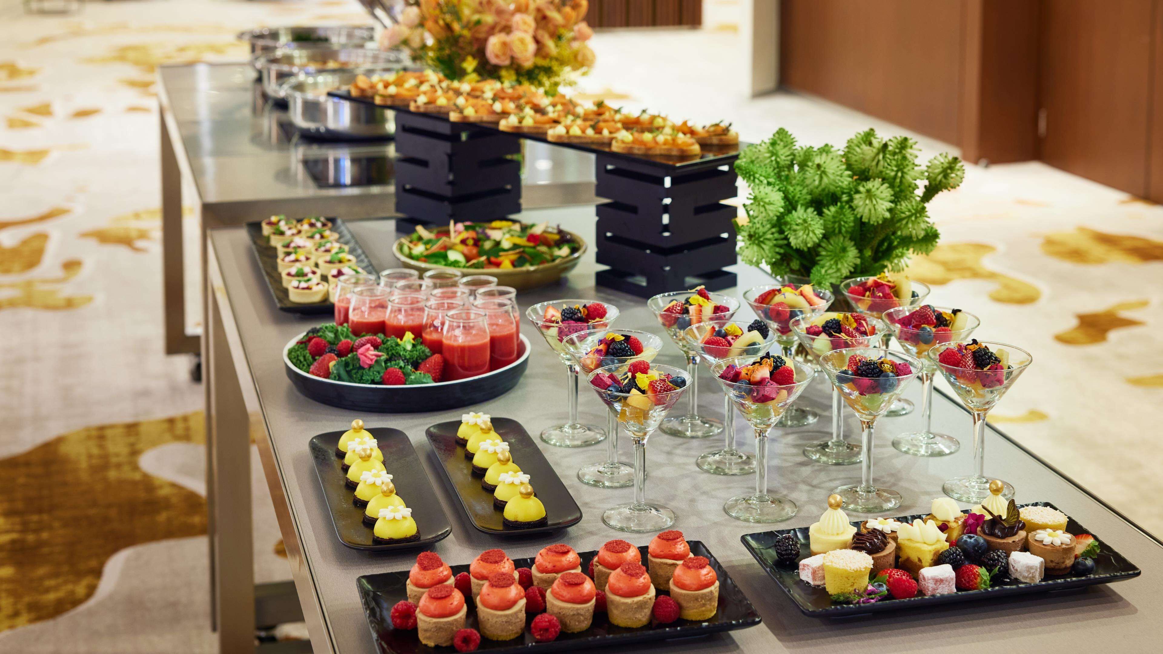 Uma mesa de buffet com diversos alimentos, incluindo frutas, sobremesas e bebidas.