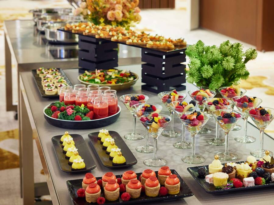 Uma mesa de buffet com diversos alimentos, incluindo frutas, sobremesas e bebidas.