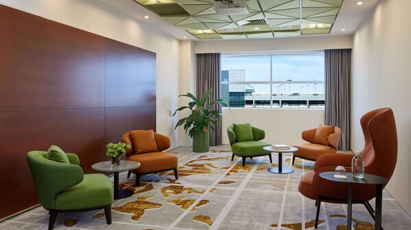 A modern lounge area with leather and green upholstered chairs, a large potted plant, and a view of the airport in the background.