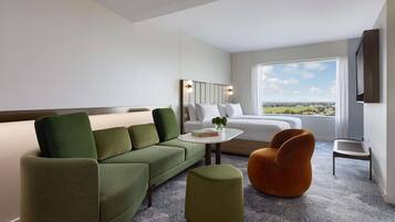 Una habitación de hotel moderna con un sofá verde, un sillón naranja pequeño, una mesa de centro blanca con un libro y una cama con ropa de cama blanca.