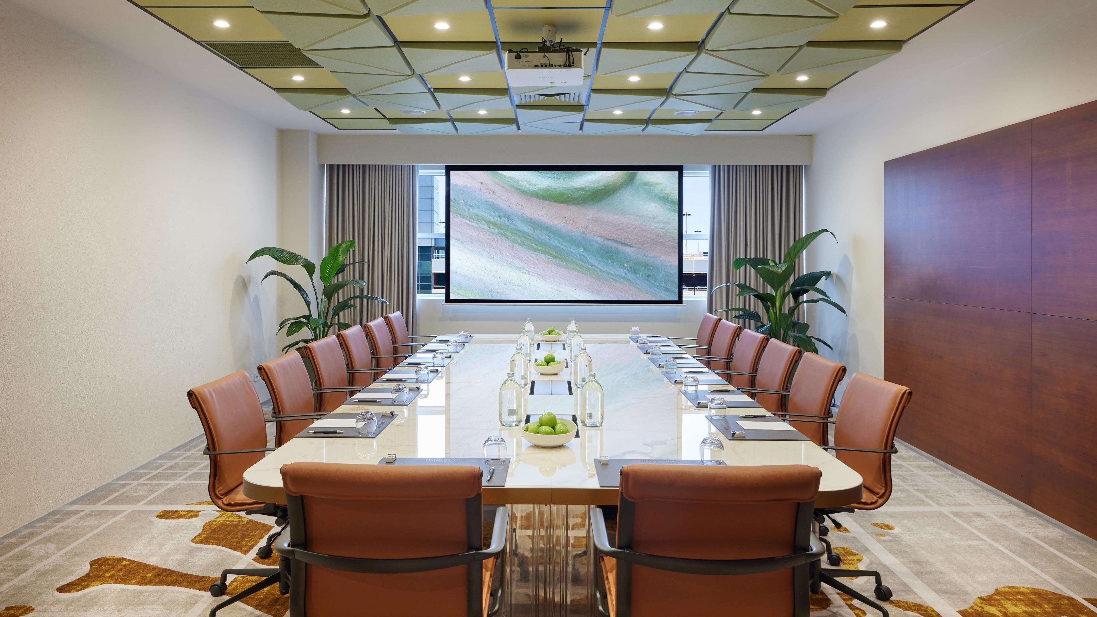 Une salle de conférence avec une longue table, des fauteuils en cuir marron, un grand écran et des plantes en pot.