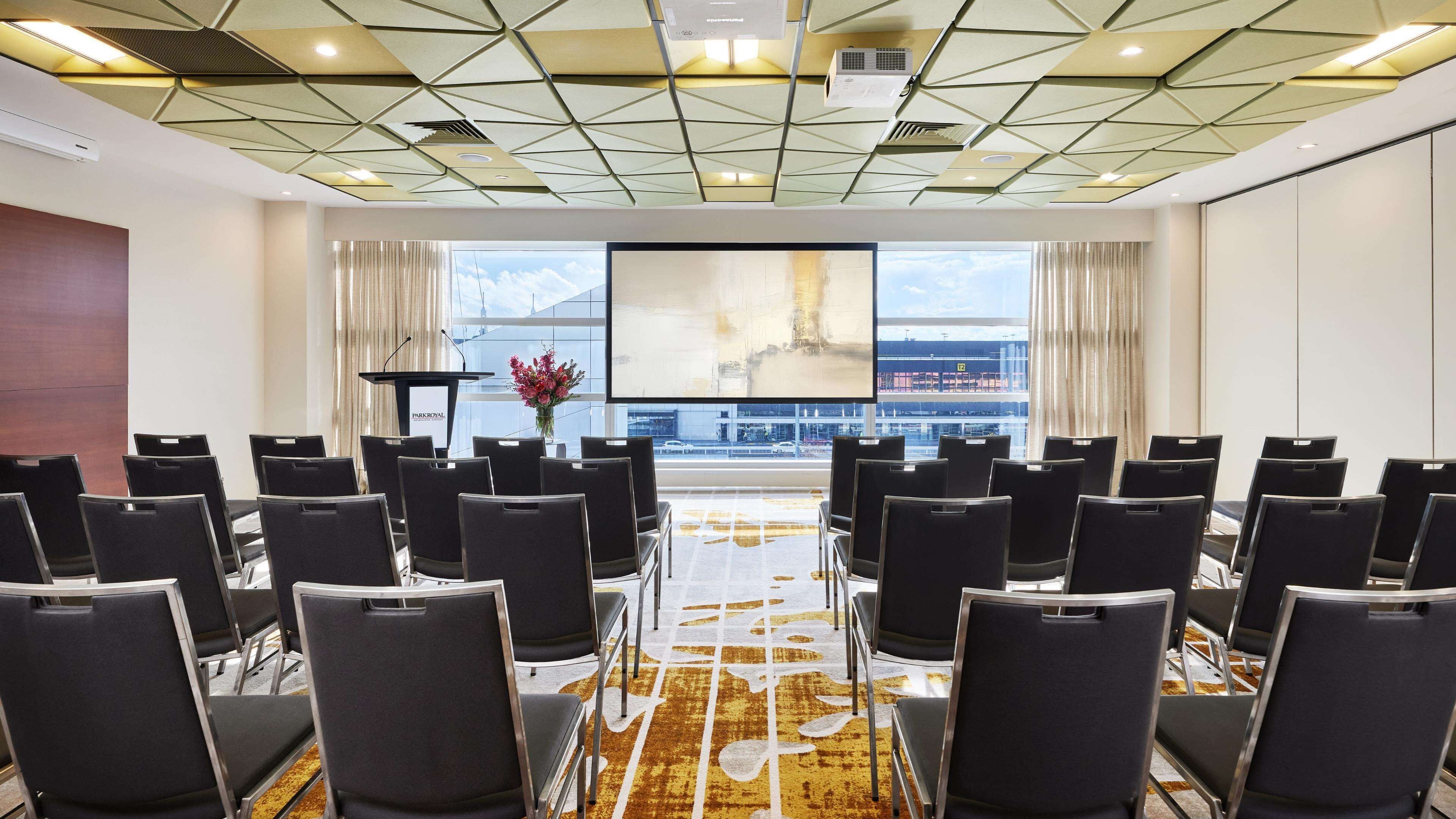 A conference room with a modern ceiling design, a large screen, and a view of the airport in the background.