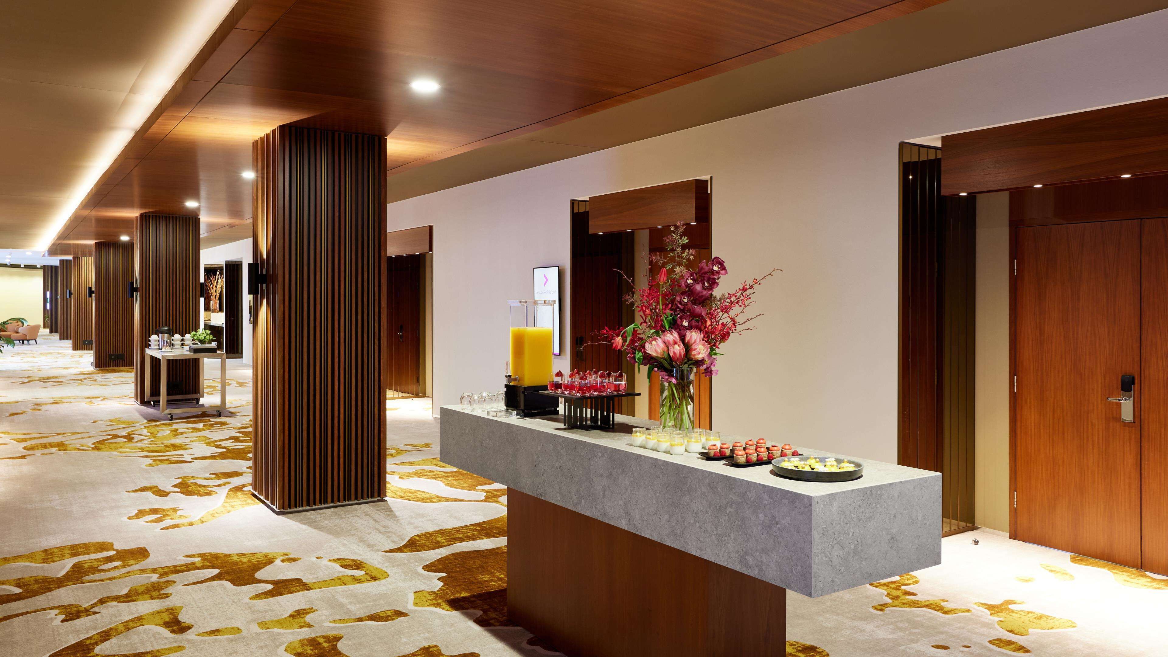 A hotel reception area with a long corridor, wooden columns, a reception desk with a vase of flowers, and a wall-mounted clock.