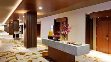 A hotel reception area with a long corridor, wooden columns, a reception desk with a vase of flowers, and a wall-mounted clock.