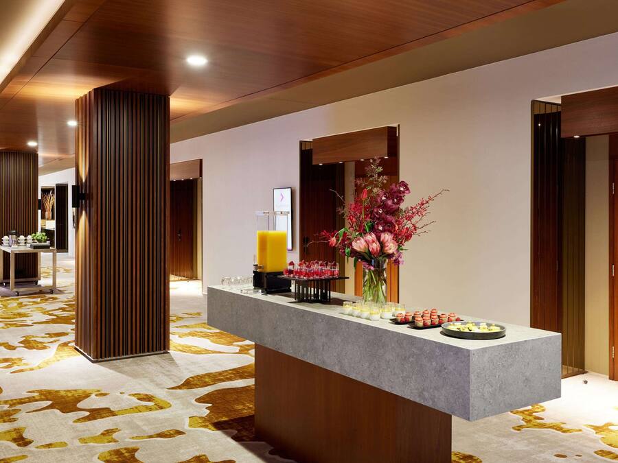 A hotel reception area with a long corridor, wooden columns, a reception desk with a vase of flowers, and a wall-mounted clock.