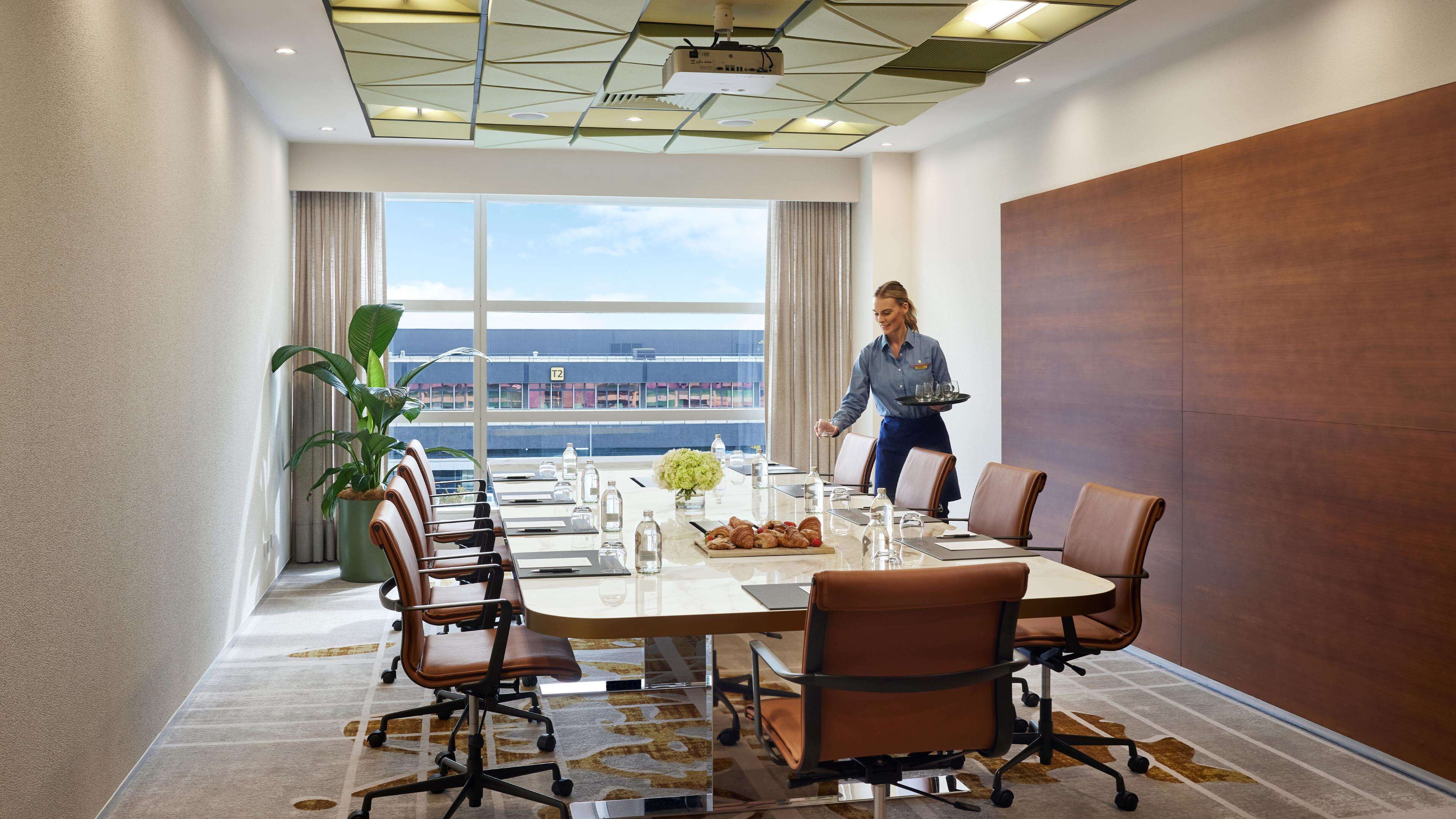 Un salón de conferencias con una mesa grande, sillas, una planta y vistas de un edificio a través de las ventanas.