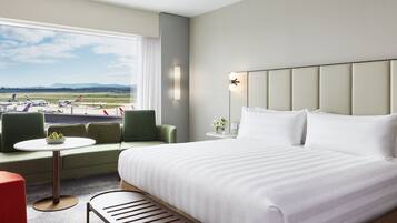 A hotel room with a large bed, a small table, a bench, and a view of an airport.
