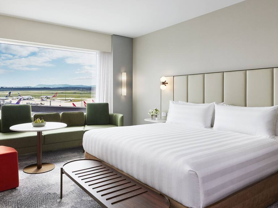 A hotel room with a large bed, a small table, a bench, and a view of an airport.