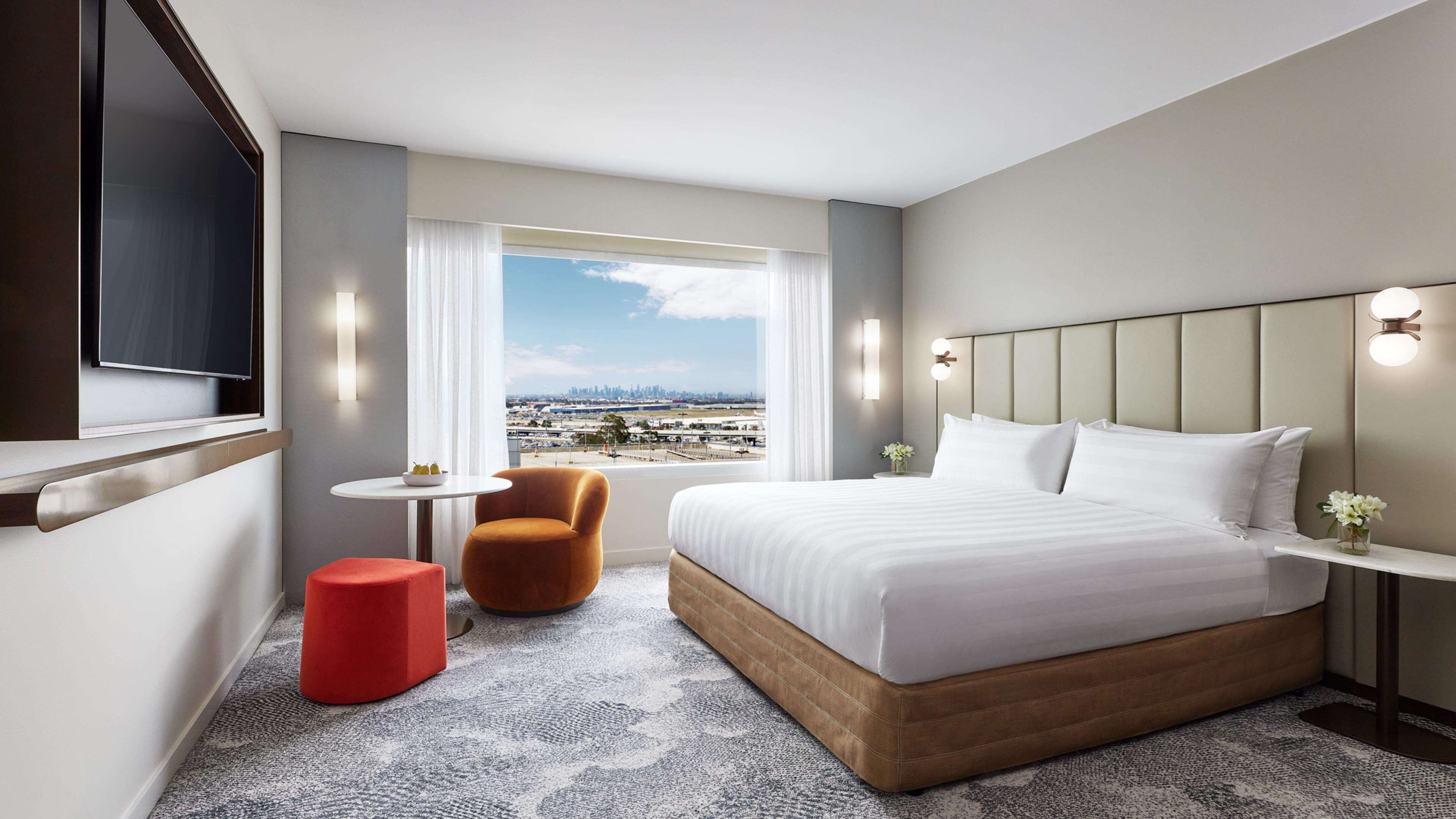 Quarto de hotel com uma cama grande, um banquinho vermelho, uma pequena mesa redonda e vista para a cidade através de uma janela ampla.