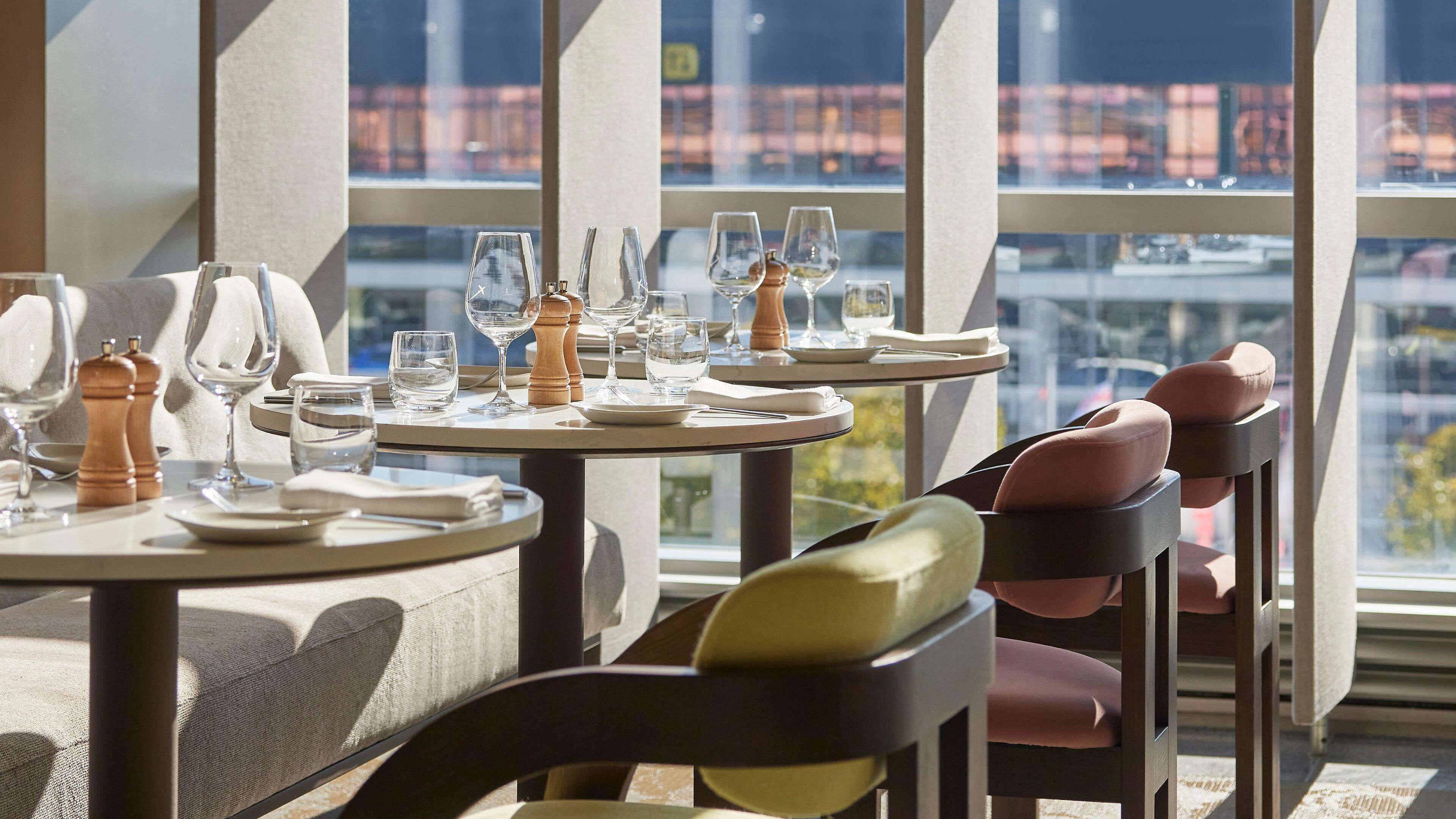 A dining table set with plates, glasses, and cutlery, with a city view through large windows.