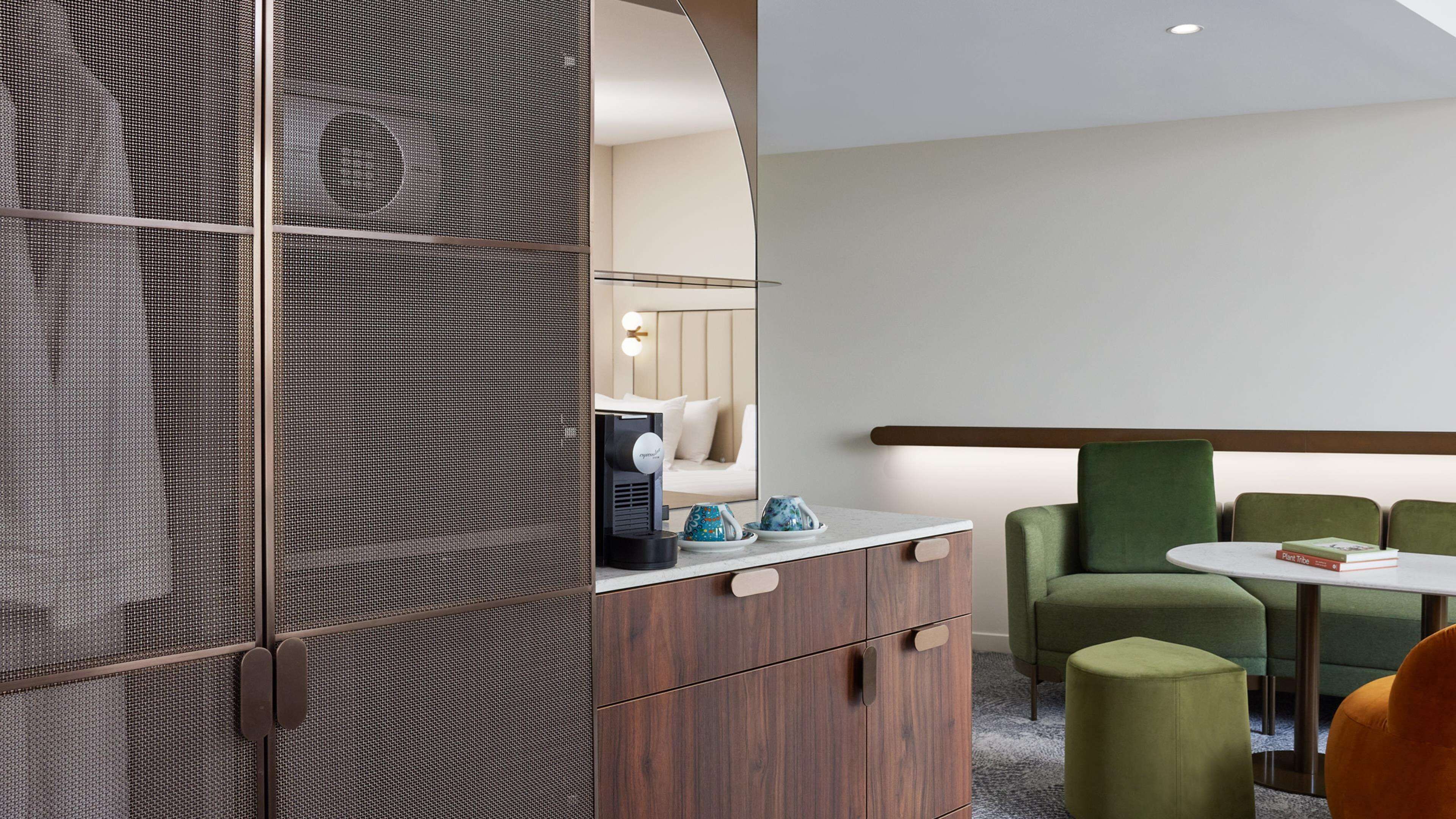 A modern hotel room with a wooden wardrobe, a small table with a coffee machine, a green armchair, and a round table with a book.