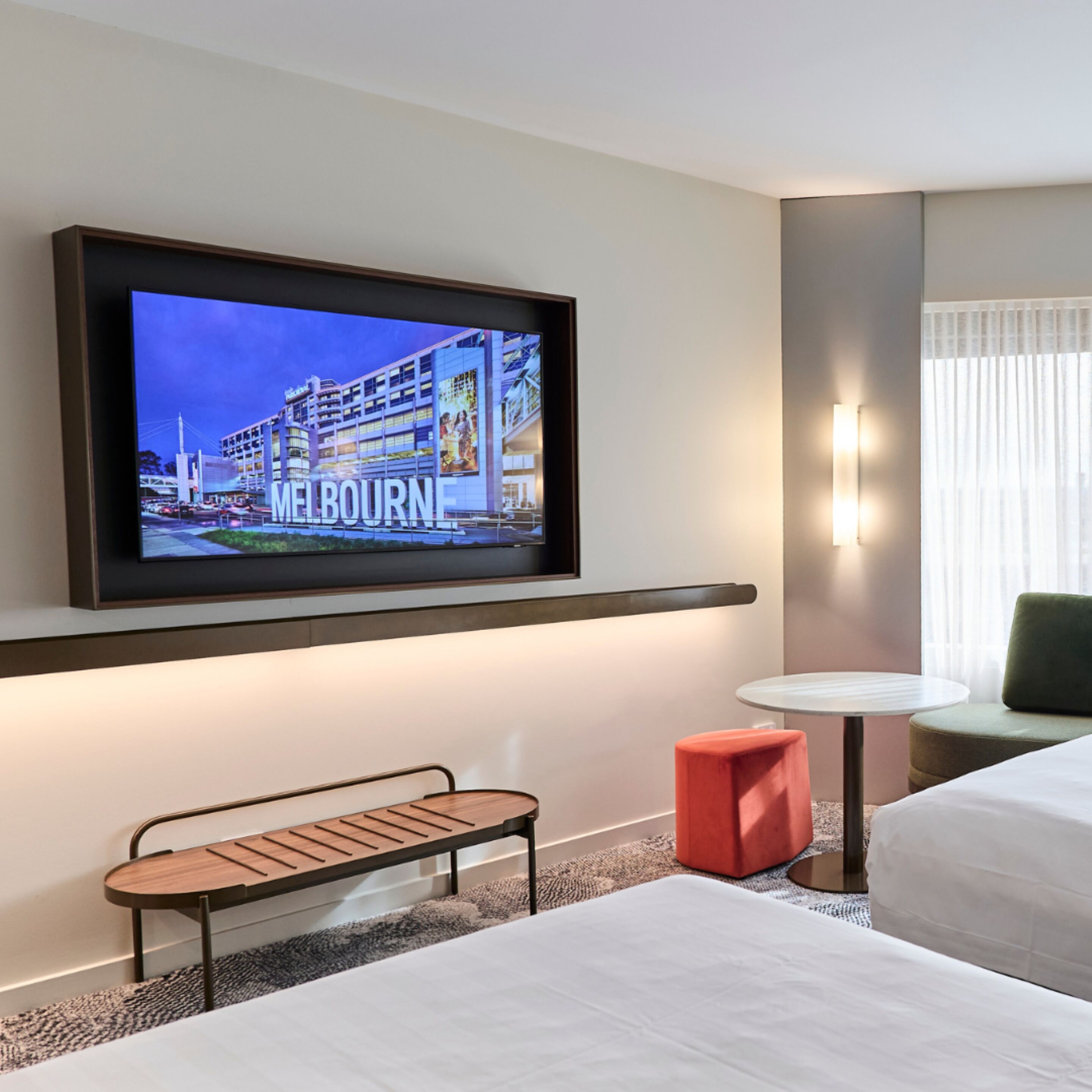 A hotel room with a television displaying a Melbourne cityscape, a small table, a bench, a red ottoman, and a bed.