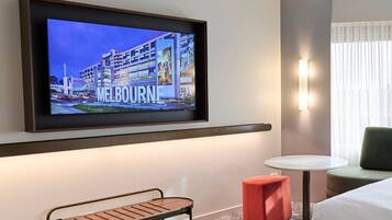 A hotel room with a television displaying a Melbourne cityscape, a small table, a bench, a red ottoman, and a bed.