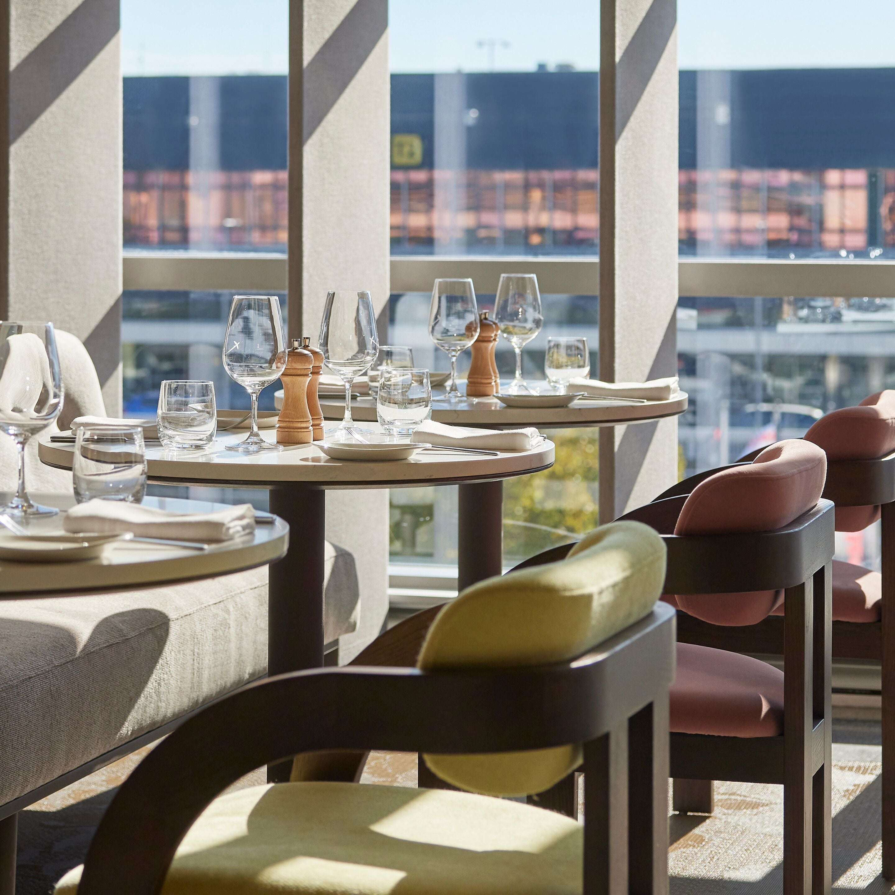 A dining area with tables set for a meal, chairs with cushions, and a view of a cityscape through large windows.