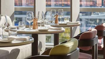A dining area with tables set for a meal, chairs with cushions, and a view of a cityscape through large windows.