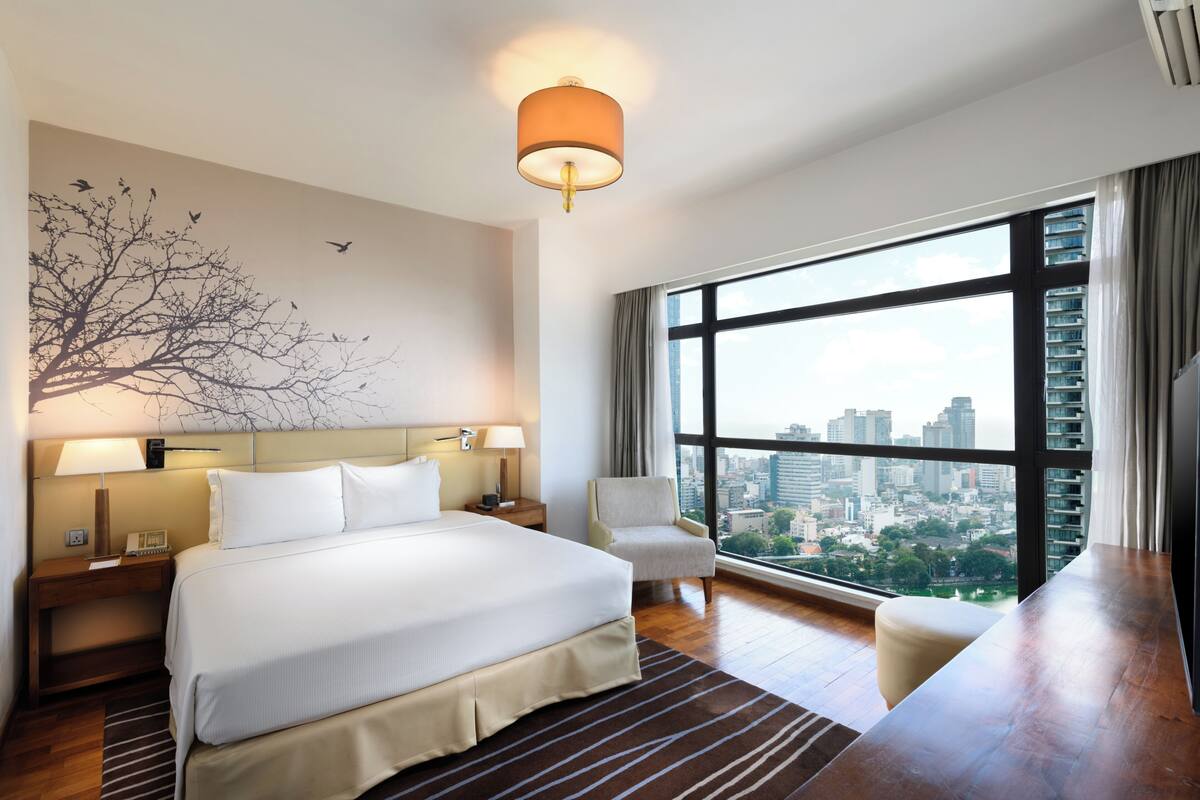 A hotel room with a large bed, two bedside tables, a chair, a view of the city, and a tree mural on the wall.