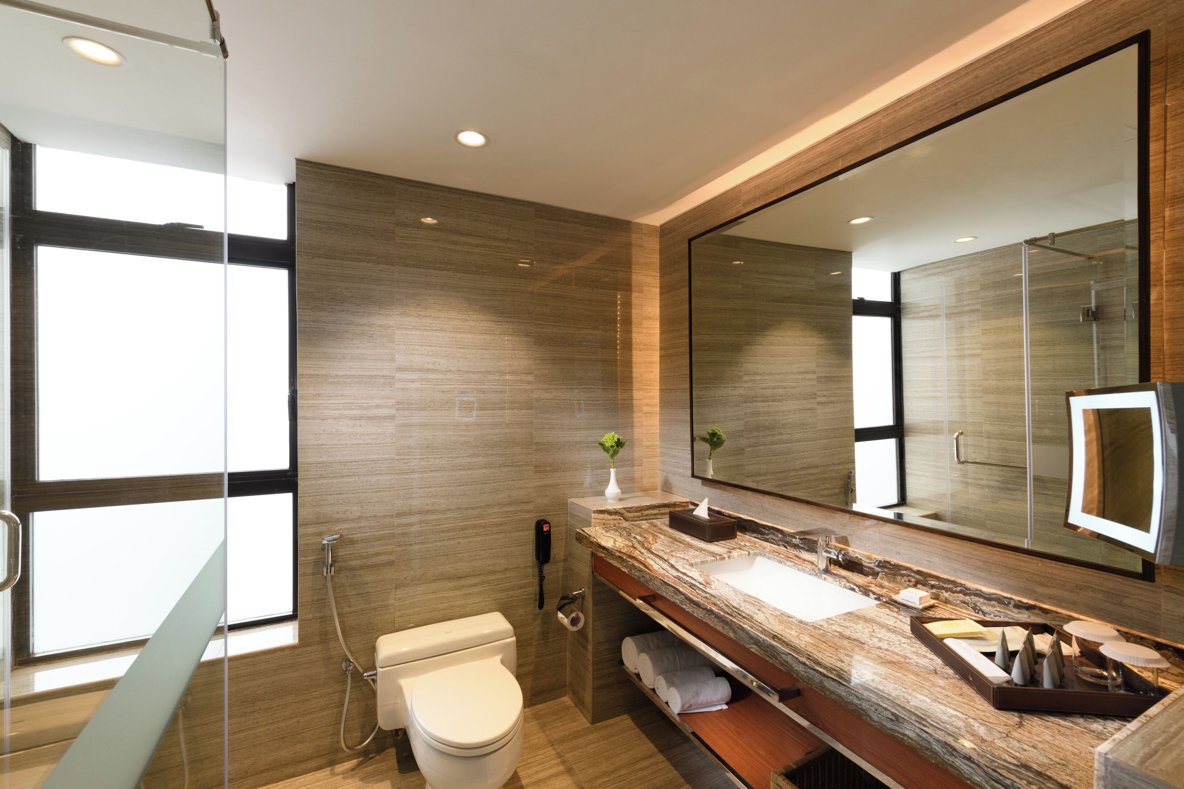 A modern bathroom with a large mirror, a marble countertop, a toilet, and a glass shower enclosure.