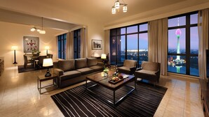 A spacious living room with a large window offering a city view, a modern sofa set, a coffee table, and a dining area with a lamp.