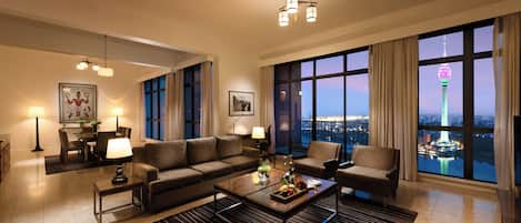 Un salón amplio con un gran ventanal que ofrece vistas a la ciudad, un conjunto de sofás modernos, una mesa de centro y un comedor con lámpara.