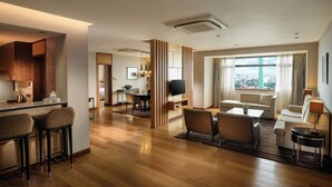 A modern hotel room with a kitchen area, a dining space, a living area with a sofa and TV, and a large window offering a city view.
