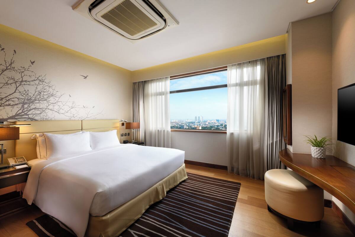 A hotel room with a large bed, a desk, a chair, a television, a window with curtains, and a city view.