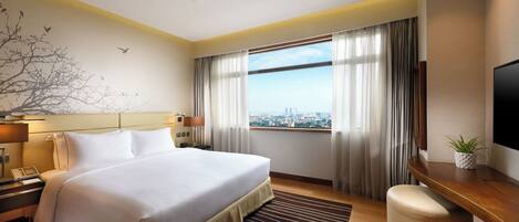 A hotel room with a large bed, a desk, a chair, a television, a window with curtains, and a city view.