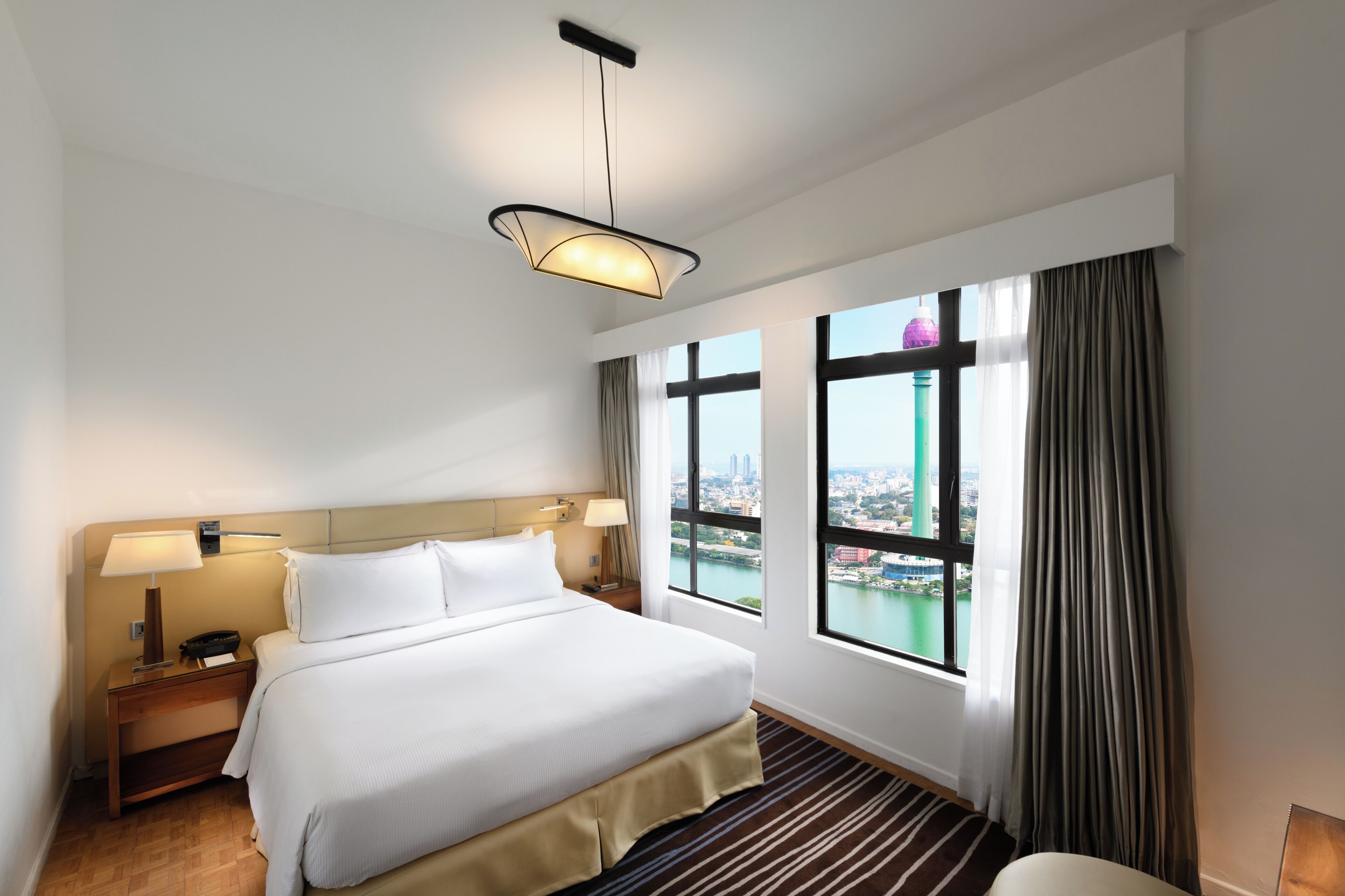 Habitación de hotel con cama, mesitas de noche, lámpara, vista panorámica de la ciudad a través de amplias ventanas y una lámpara colgante.