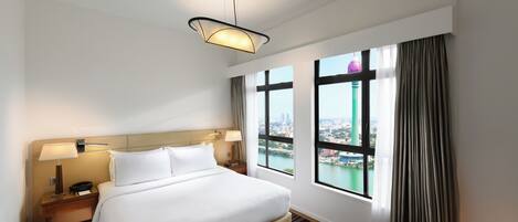 Habitación de hotel con cama, mesitas de noche, lámpara, vista panorámica de la ciudad a través de amplias ventanas y una lámpara colgante.