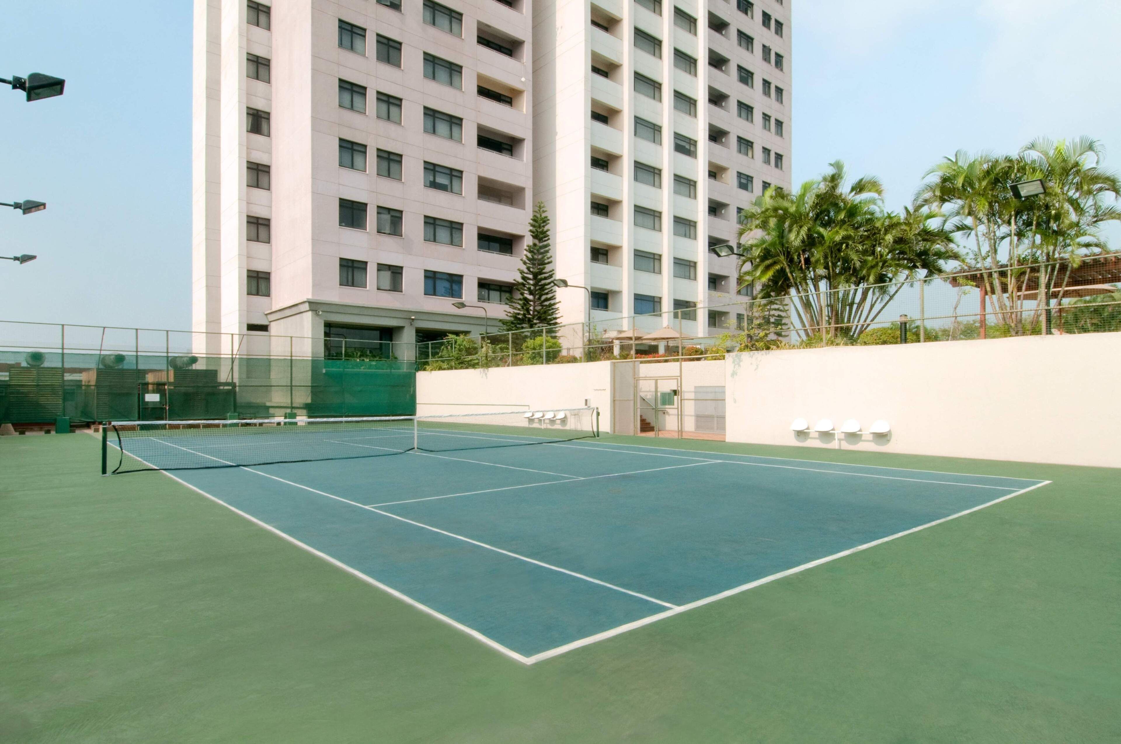 An outdoor tennis court with a net, surrounded by a low fence, and a tall building in the background.