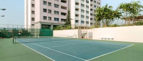 An outdoor tennis court with a net, surrounded by a low fence, and a tall building in the background.