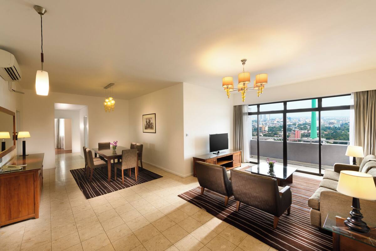 A modern hotel room with a dining area, a living area with a TV, a large window with a city view, and a chandelier.