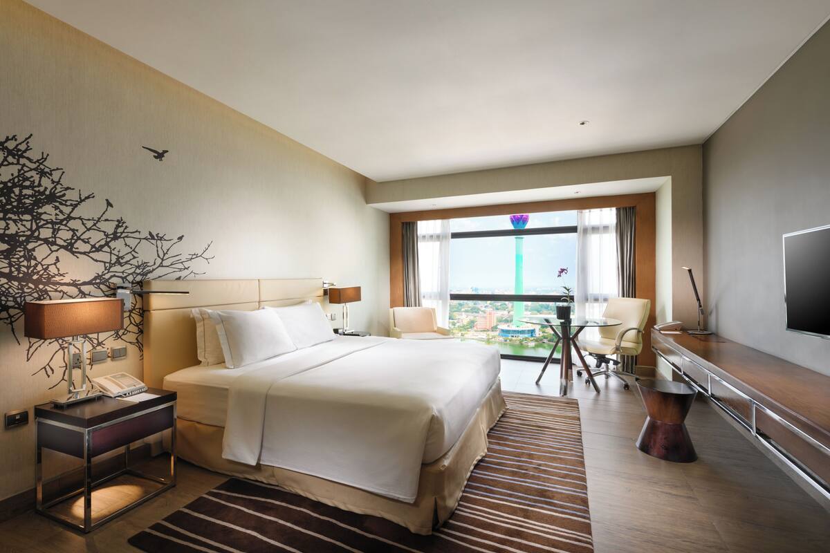A hotel room with a bed, bedside tables, a TV, a desk, a chair, and a view of the city.