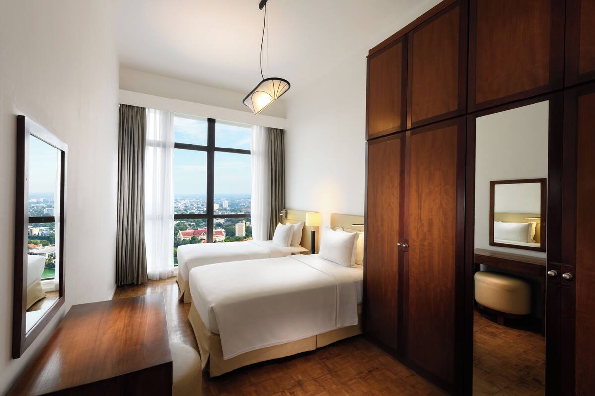 A hotel room with two beds, a wooden wardrobe, a bedside table, a mirror, and a view of the city.