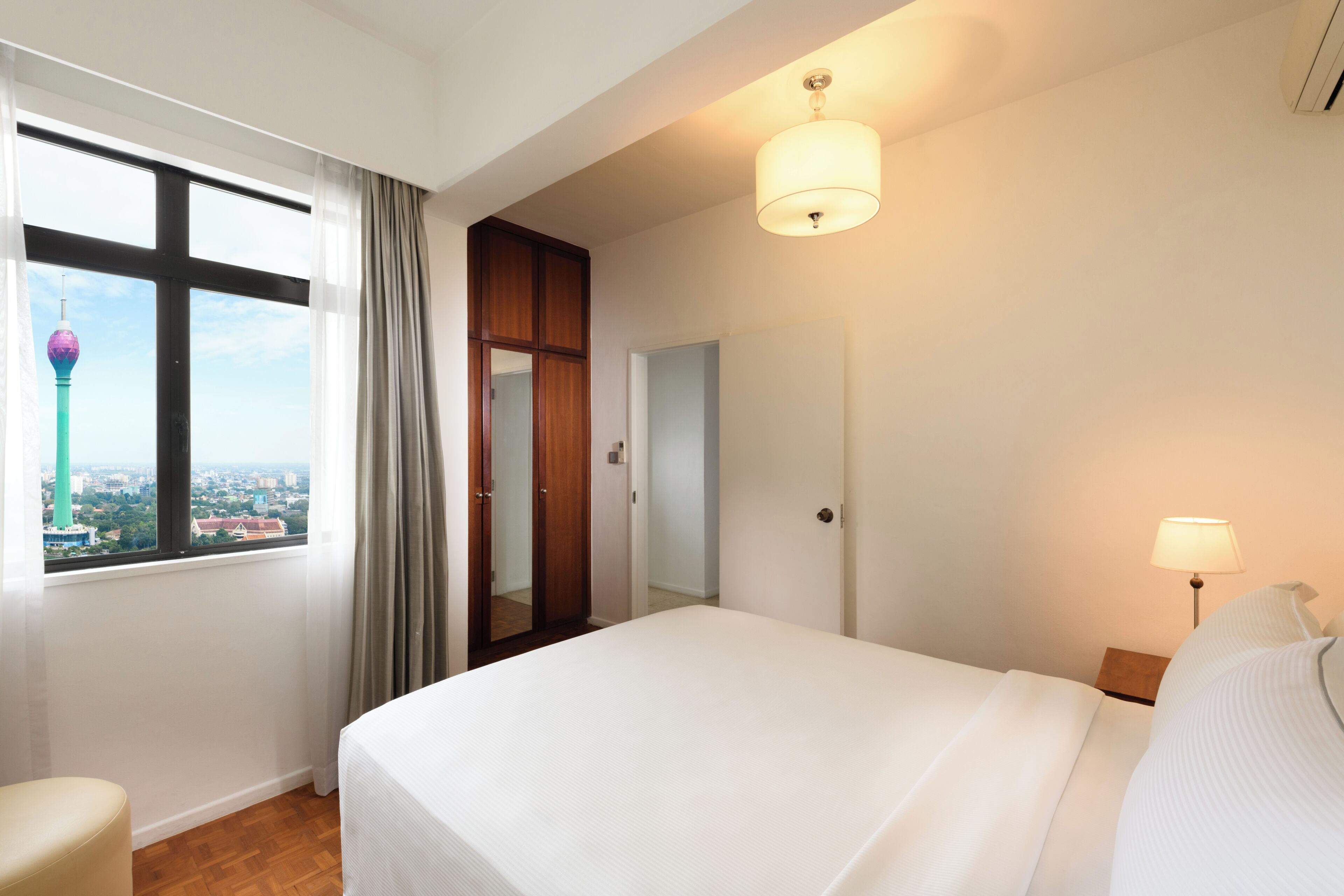 Ein Hotelzimmer mit Bett, Nachttisch, Lampe, Fenster mit Blick auf einen Turm und einer Tür zu einem anderen Raum.