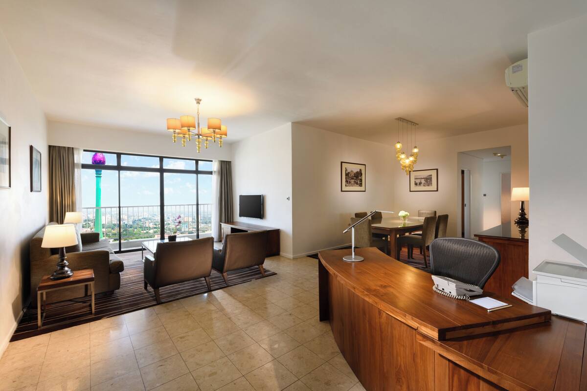 A hotel room with a desk, chairs, a television, a chandelier, and a large window with a view.