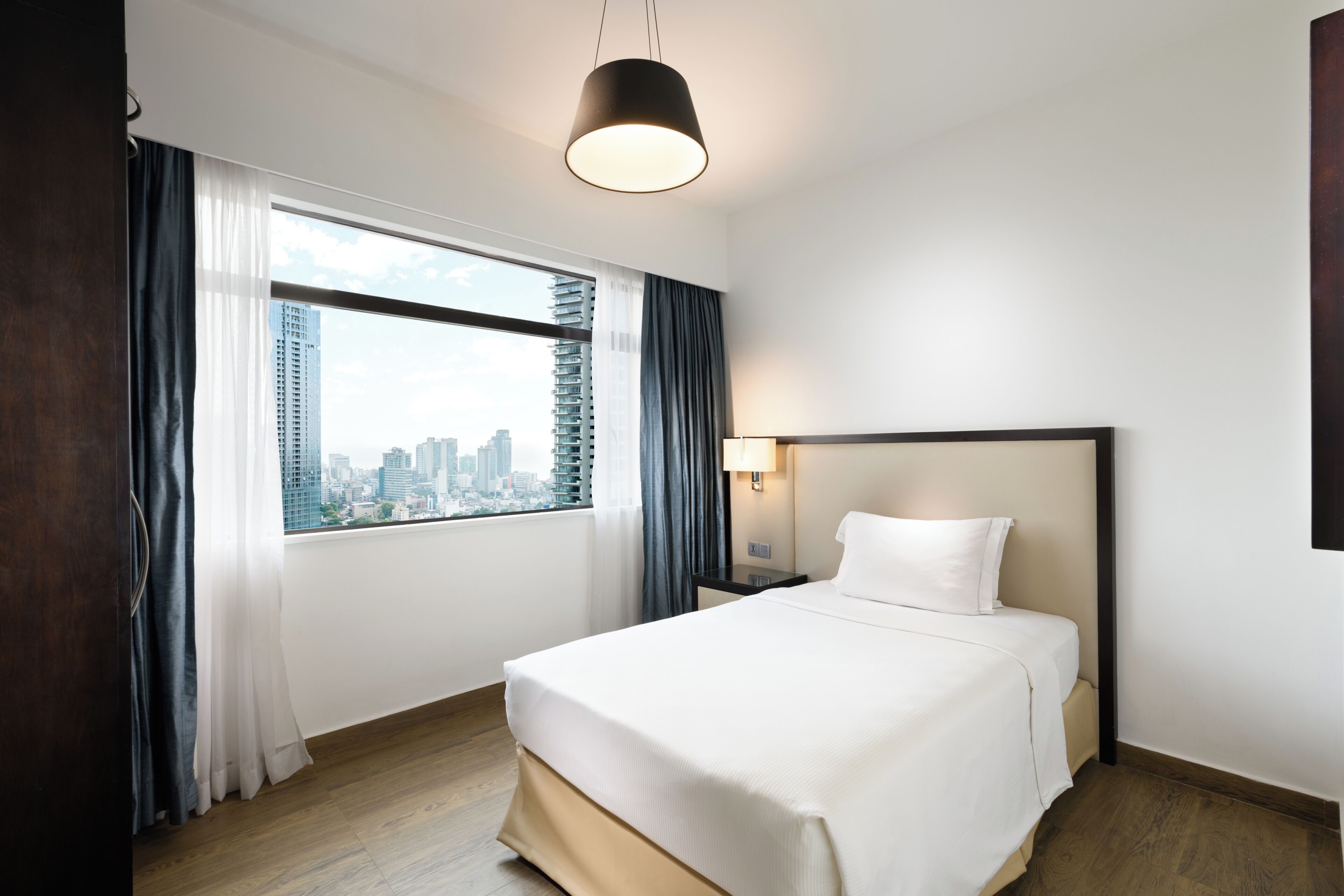 A hotel room with a large bed, a nightstand, a lamp, a window with curtains, and a city view.