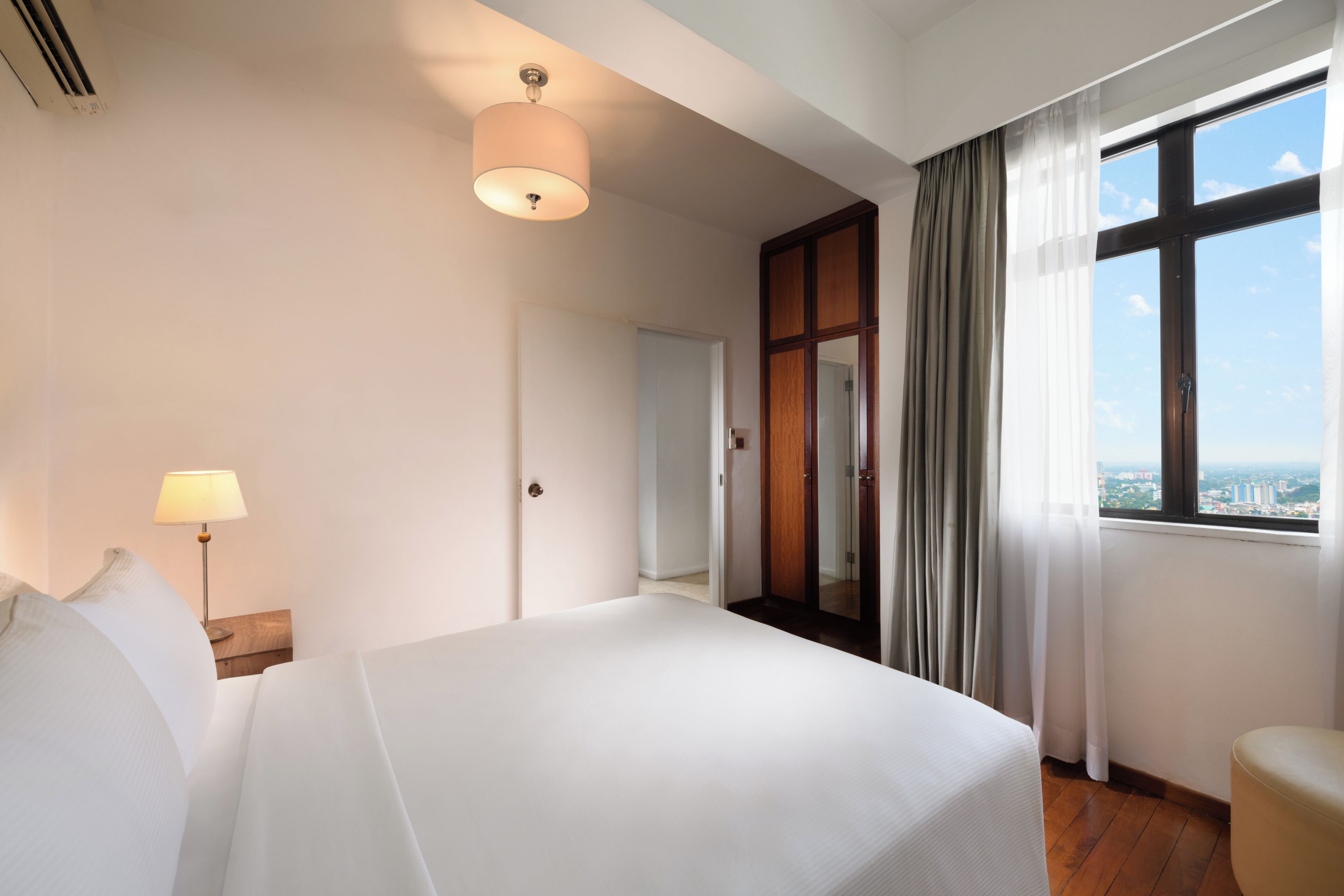 A hotel room with a bed, a nightstand, a lamp, a window with curtains, and a view of the city.