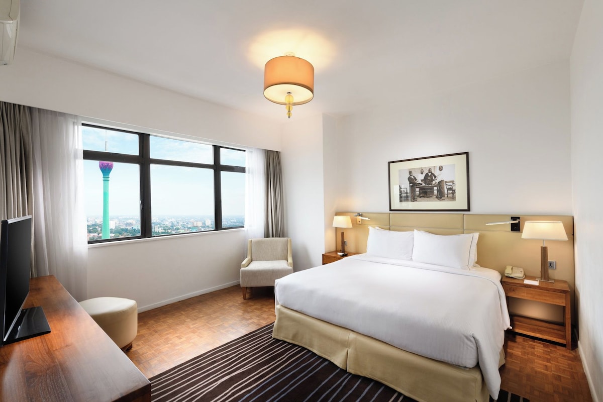 A hotel room with a large bed, a desk, a chair, a nightstand, a lamp, a framed picture on the wall, and a window with a view of a tower.