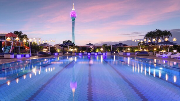 A swimming pool with lounge chairs and umbrellas, a tower with a lighted top, and a sunset sky.