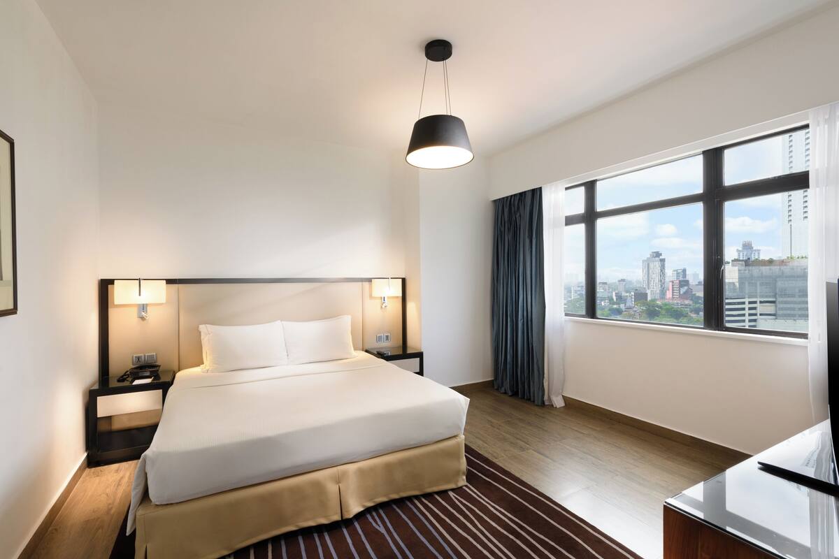 A hotel room with a large bed, bedside lamps, a desk, and a view of the city through large windows.