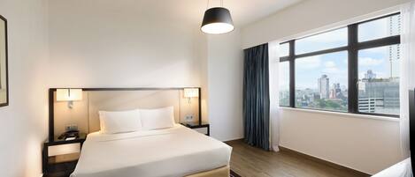 Quarto de hotel com uma cama grande, luminárias de cabeceira, uma escrivaninha e vista para a cidade através de amplas janelas.