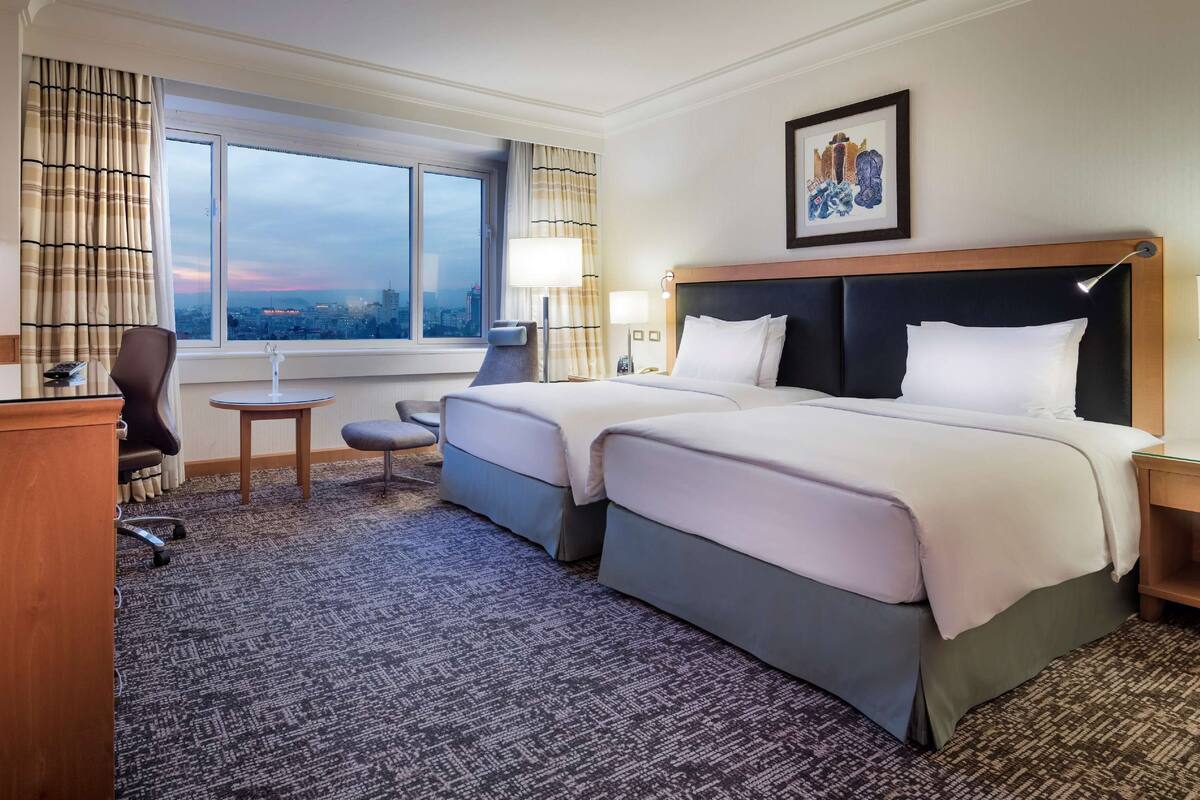 A hotel room with a large bed, a desk with a computer, a chair, a small table, and a window with a city view.